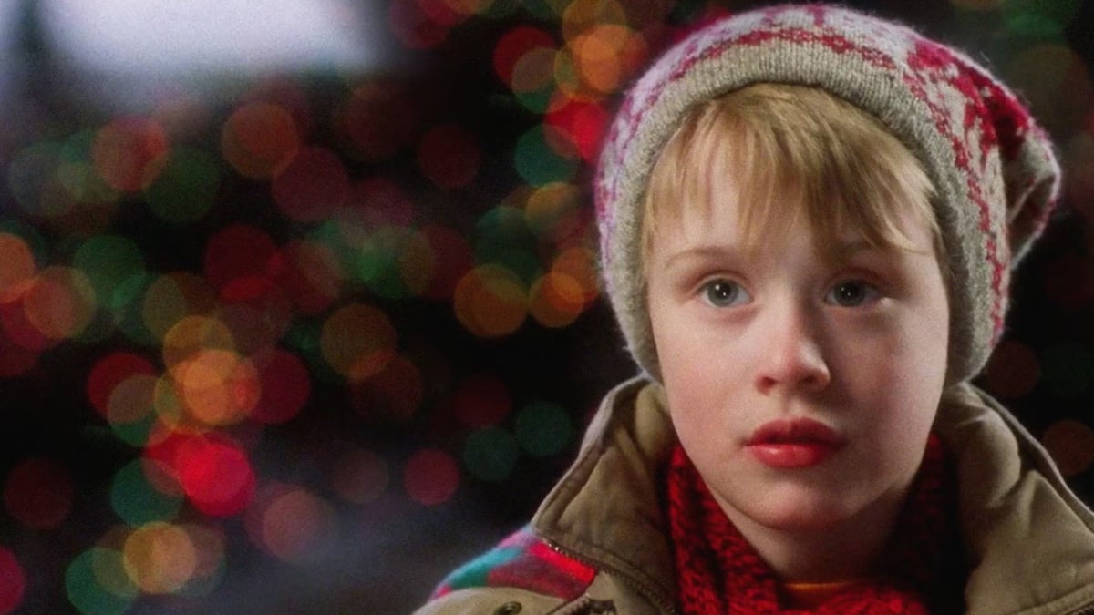 A young child wearing a knit winter hat and coat stands in front of colorful, out-of-focus holiday lights, looking thoughtful.