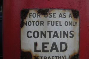 A weathered metal sign on a red surface reads "FOR USE AS A MOTOR FUEL ONLY CONTAINS LEAD (TETRAETHYL)," with rust visible around the edges of the sign.