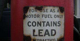 A weathered metal sign on a red surface reads "FOR USE AS A MOTOR FUEL ONLY CONTAINS LEAD (TETRAETHYL)," with rust visible around the edges of the sign.