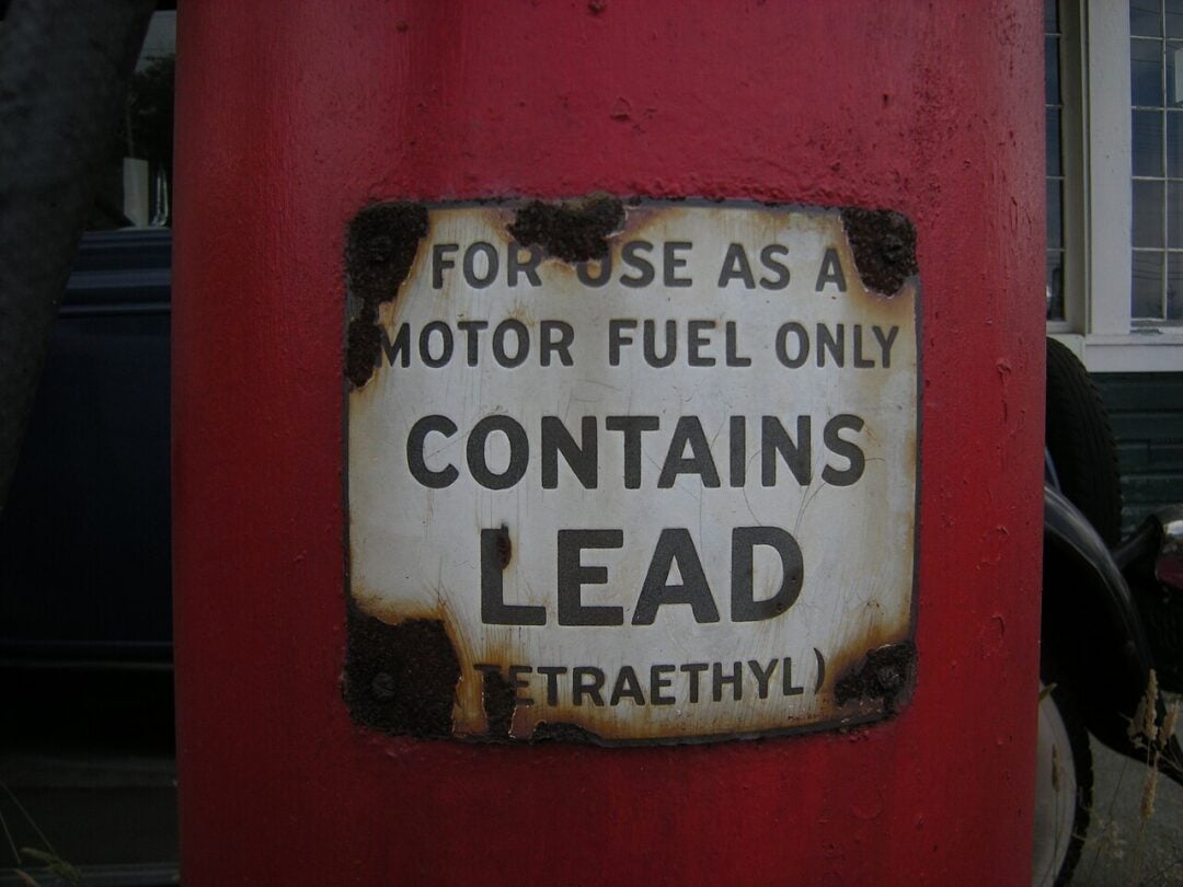 A weathered sign on a red surface reads: "FOR USE AS A MOTOR FUEL ONLY CONTAINS LEAD (TETRAETHYL)." The metal sign is rusted at the corners.