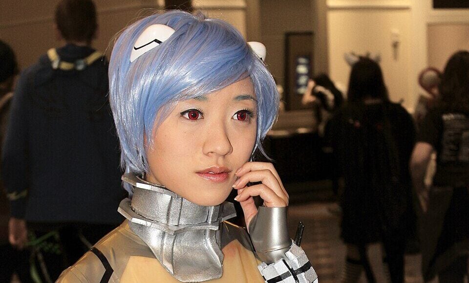 A person with short blue hair and a futuristic, translucent bodysuit labeled "TEST" cosplays at a convention, standing indoors with other attendees visible in the background.