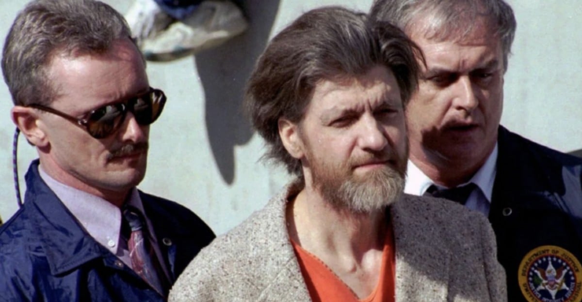 A bearded man in an orange prison jumpsuit and tan jacket is escorted outdoors by two law enforcement officers, one wearing sunglasses and the other in a dark jacket with an official seal.