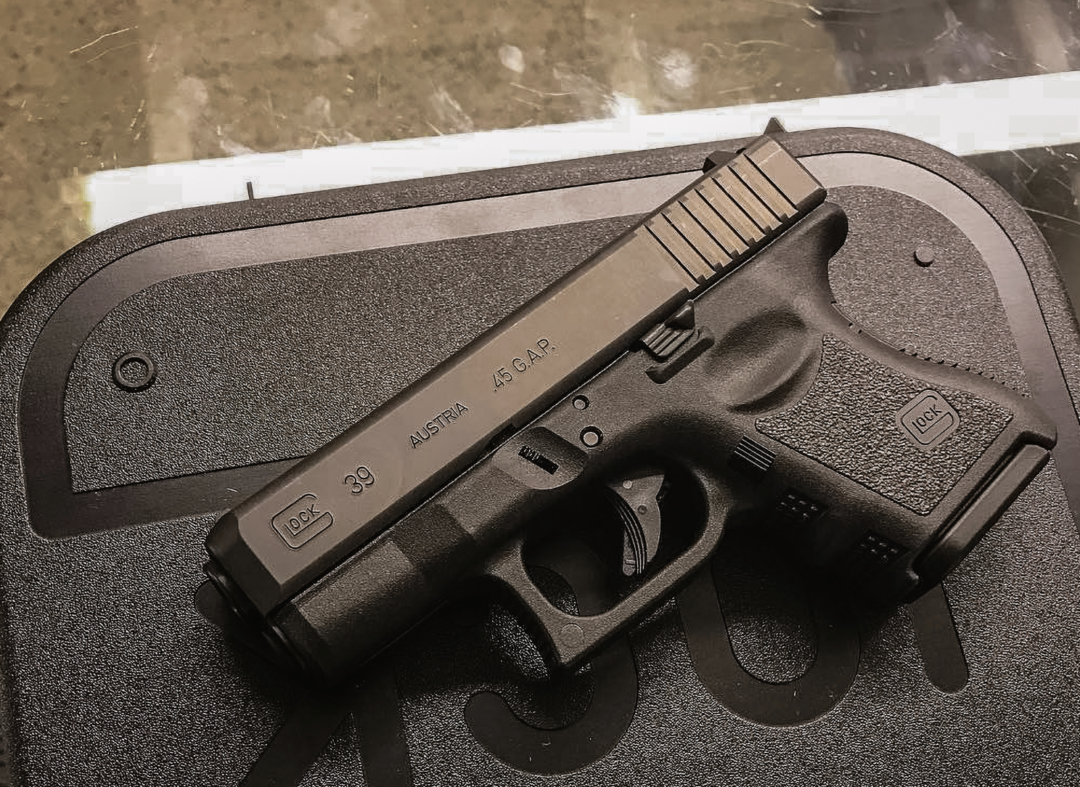 A black Glock 39 pistol is resting on a textured black case. The pistol has markings indicating it is chambered in .45 GAP and manufactured in Austria.