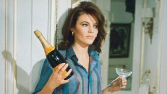 A woman with long brown hair, wearing a striped shirt, holds a bottle of champagne in one hand and an empty glass in the other, standing indoors near a white wall.