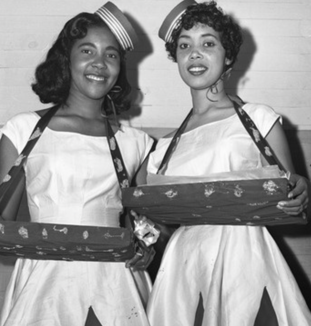 Two women in matching white dresses and striped hats smile at the camera. They each wear a tray around their necks, possibly for serving or selling items, giving a festive or promotional appearance.