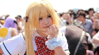 A person in a school uniform cosplay with long blonde hair, a red plaid bow, and a blue scrunchie, stands thoughtfully in a crowded outdoor event, holding strands of hair near their mouth.