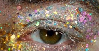 A close-up of a person's eye with brown iris, surrounded by eyelids and skin covered in colorful, sparkling glitter in shades of gold, pink, blue, and silver. The eyebrow is visible at the top.