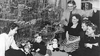 A vintage black-and-white photo shows a family of six gathered around a Christmas tree, smiling. A small child sits on the floor with toys, while four adults and an older child sit or kneel around the tree.