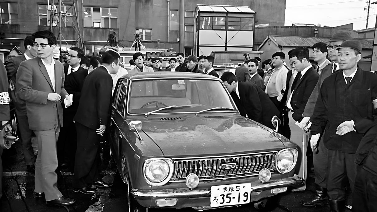 A crowd of men in suits and uniforms gathers around a compact car in a parking lot, some closely inspecting it, with buildings visible in the background. The photo appears to be from mid-20th century Japan.