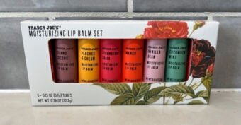 A set of six Trader Joe's moisturizing lip balms in a floral box. Flavors include Island Coconut, Peaches & Cream, Strawberry, Mango, Vanilla Bean, and Cucumber Mint. The package is displayed against a gray tile background.