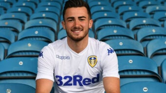 A smiling man in a white Leeds United football jersey sits in an empty stadium with blue seats.
