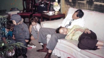 A man sits on a couch with a child asleep on his lap, both resting. Nearby, two children play on the floor while another sits quietly. The scene appears cozy and domestic, with a partially visible Christmas tree in the foreground.