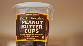 A clear plastic container labeled &ldquo;Trader Joe&rsquo;s Dark Chocolate Peanut Butter Cups&rdquo; with yellow and brown colors, showing peanut butter cups inside. The label notes they are creamy peanut butter covered in dark chocolate.