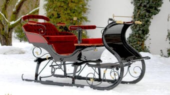 A vintage, black and red horse-drawn sleigh with elegant gold details sits on snow, surrounded by trees and ivy-covered walls. The sleigh features plush red velvet seats and ornate metalwork.
