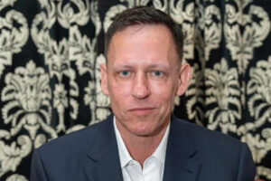 A man in a dark suit and white shirt sits in front of a black and white ornate patterned curtain, looking at the camera with a neutral expression.