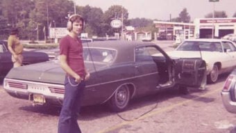 A young man wearing headphones stands beside a vintage green car with the driver&rsquo;s door open in a parking lot; there are other parked cars and a gas station in the background.