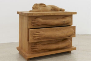 A wooden chest of drawers features sculpted arms and hands on each drawer front, with a carved rabbit lying on top. The chest sits on a plain floor against a white wall.
