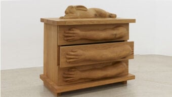 A wooden chest of drawers features sculpted arms and hands on each drawer front, with a carved rabbit lying on top. The chest sits on a plain floor against a white wall.