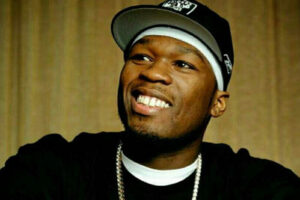 A man wearing a black baseball cap, black shirt, white headband, and a silver chain necklace smiles while looking slightly to the side. The background is blurred and neutral-toned.