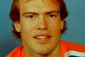 A young man with light brown hair is wearing an orange sports jersey and looking at the camera, with a blue background behind him.