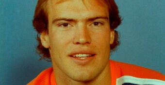 A young man with light brown hair is wearing an orange sports jersey and looking at the camera, with a blue background behind him.