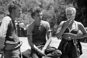 Three young adults stand outdoors in casual 1950s attire; two men talk while a woman in a bikini top and jacket smokes a cigarette. Trees and more people are visible in the background.