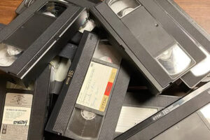 A pile of VHS tapes is stacked on a wooden surface. Some tapes have handwritten labels, while others are blank or have printed stickers. The tapes appear old and slightly worn.
