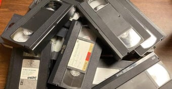 A pile of VHS tapes is stacked on a wooden surface. Some tapes have handwritten labels, while others are blank or have printed stickers. The tapes appear old and slightly worn.