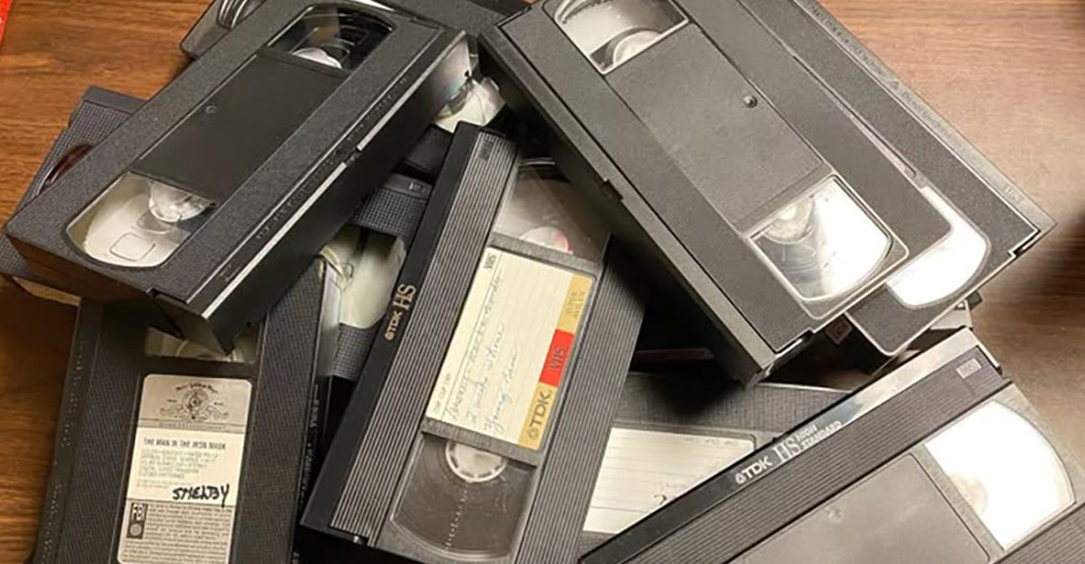 A pile of VHS tapes is stacked on a wooden surface. Some tapes have handwritten labels, while others are blank or have printed stickers. The tapes appear old and slightly worn.