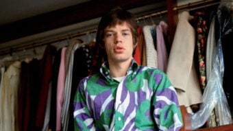 A young man with straight brown hair wears a bold, colorful shirt with green and purple patterns, standing in front of a rack of assorted clothes and jackets hanging in a wardrobe.