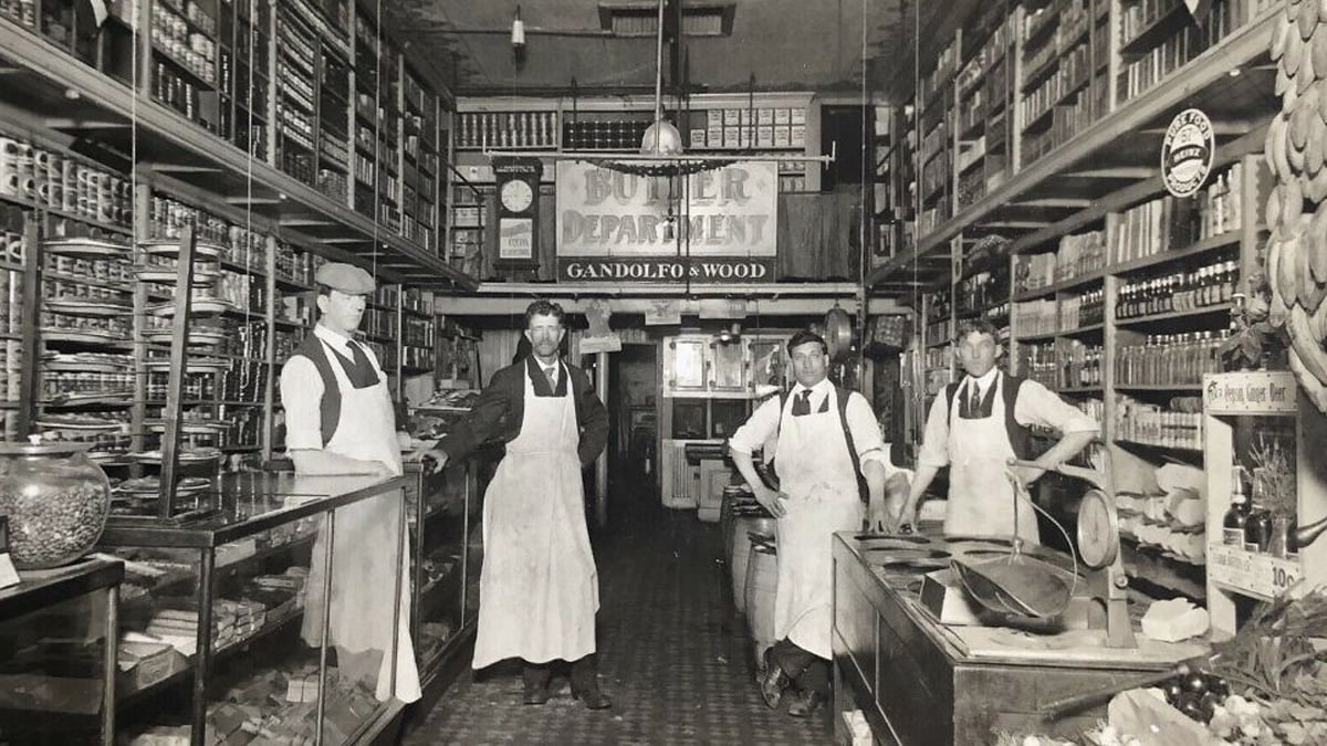 Four men in aprons stand inside an old-fashioned grocery store or butcher shop, surrounded by shelves stocked with canned goods, jars, and boxes. A “Butcher Department” sign hangs in the background.