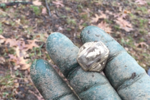 A gloved, dirt-covered hand holds a rough, metallic-looking rock or crystal, with fallen leaves and grass visible on the ground in the blurred background.