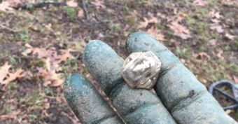A gloved, dirt-covered hand holds a rough, metallic-looking rock or crystal, with fallen leaves and grass visible on the ground in the blurred background.