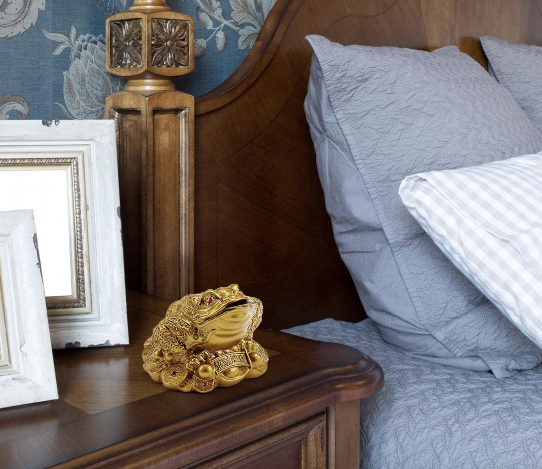 A gold three-legged frog figurine with coins sits on a wooden nightstand beside a bed with gray pillows, near framed pictures and a wooden lamp, with a floral wallpaper background.