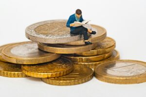 A small figurine of a person reading a book sits on a stack of large gold coins, symbolizing financial literacy or investment knowledge.