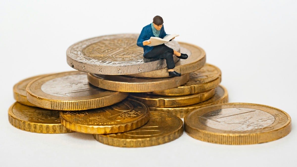 A small figurine of a person reading a book sits on a stack of large gold coins, symbolizing financial literacy or investment knowledge.