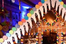 A large, colorful gingerbread house decorated with candies, icing, and chocolate hangs inside a festive room with bright lights and artificial snow on the ground. The house has a candy-covered roof and windows.