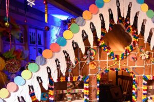 A large, colorful gingerbread house decorated with candies, icing, and chocolate hangs inside a festive room with bright lights and artificial snow on the ground. The house has a candy-covered roof and windows.