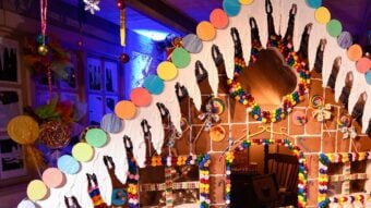 A large, colorful gingerbread house decorated with candies, icing, and chocolate hangs inside a festive room with bright lights and artificial snow on the ground. The house has a candy-covered roof and windows.