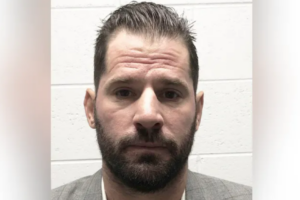 A man with short dark hair and a beard, wearing a light gray suit jacket and white shirt, stands against a white brick wall, looking directly at the camera with a neutral expression.