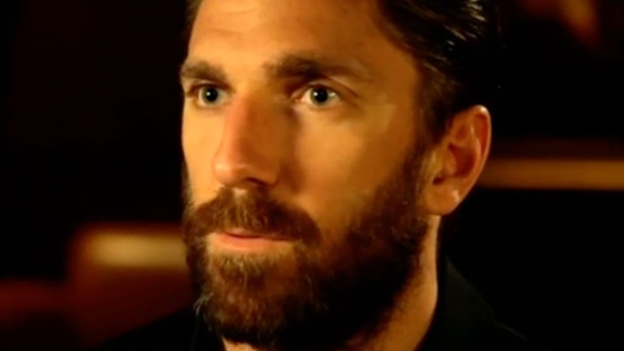 A close-up of a man with blue eyes, brown hair, and a full beard, looking slightly to the side with a neutral expression against a softly blurred dark background.