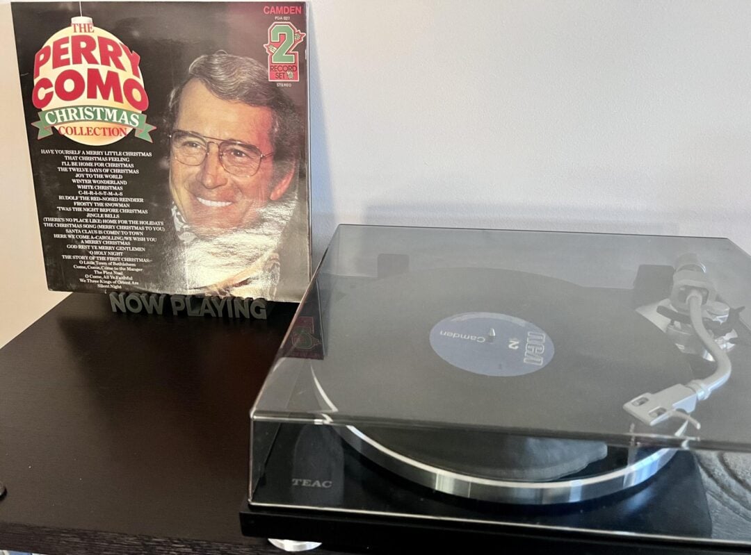 A vinyl record player spins a record, with the cover of "The Perry Como Christmas Collection" propped up behind it, featuring a smiling Perry Como and a list of songs, against a light-colored wall.