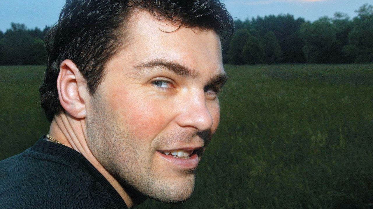A man with short dark hair and light eyes is smiling while looking over his shoulder outdoors. He stands in a grassy field with trees in the background and a cloudy sky at dusk.