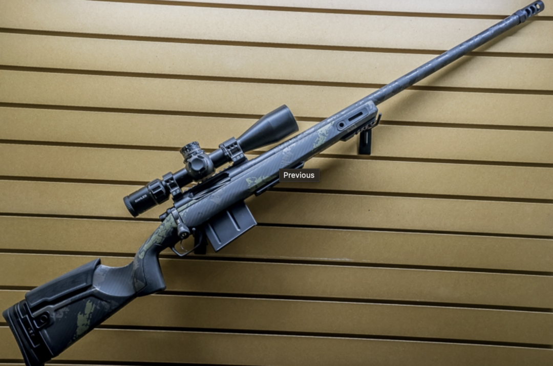 A black bolt-action sniper rifle with a mounted scope is displayed horizontally on a slatted tan wall. The rifle has a camouflaged stock and an adjustable cheek rest.