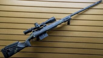 A black bolt-action sniper rifle with a scope attached is displayed horizontally on a slatted tan wall. The rifle features a camouflage pattern on the stock and a long barrel.