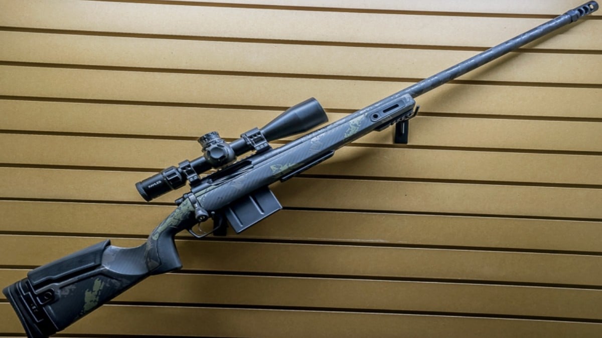 A black bolt-action sniper rifle with a scope attached is displayed horizontally on a slatted tan wall. The rifle features a camouflage pattern on the stock and a long barrel.