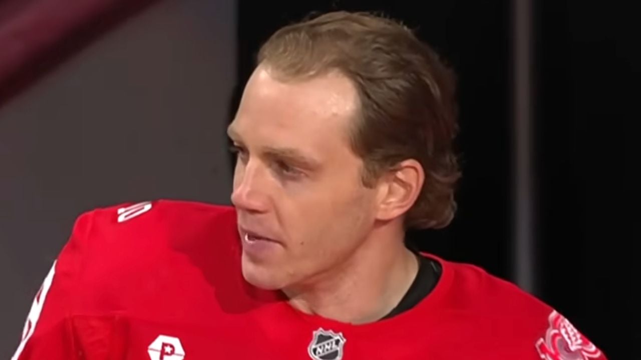 A hockey player in a red jersey looks to the side. The jersey features NHL and team logos. The background is blurred.