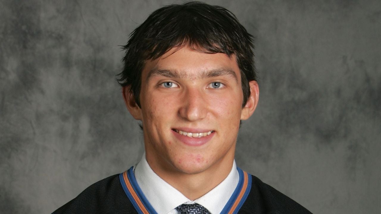 A young man with short dark hair, wearing a black jacket, white shirt, and patterned tie, smiles at the camera against a gray mottled background.