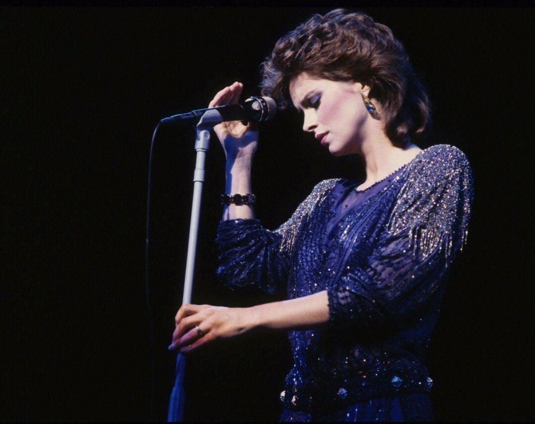 A singer in a sparkly, dark blue outfit stands at a microphone on stage, head bowed and eyes closed, with one hand on the mic stand and the other holding the mic, against a dark background.