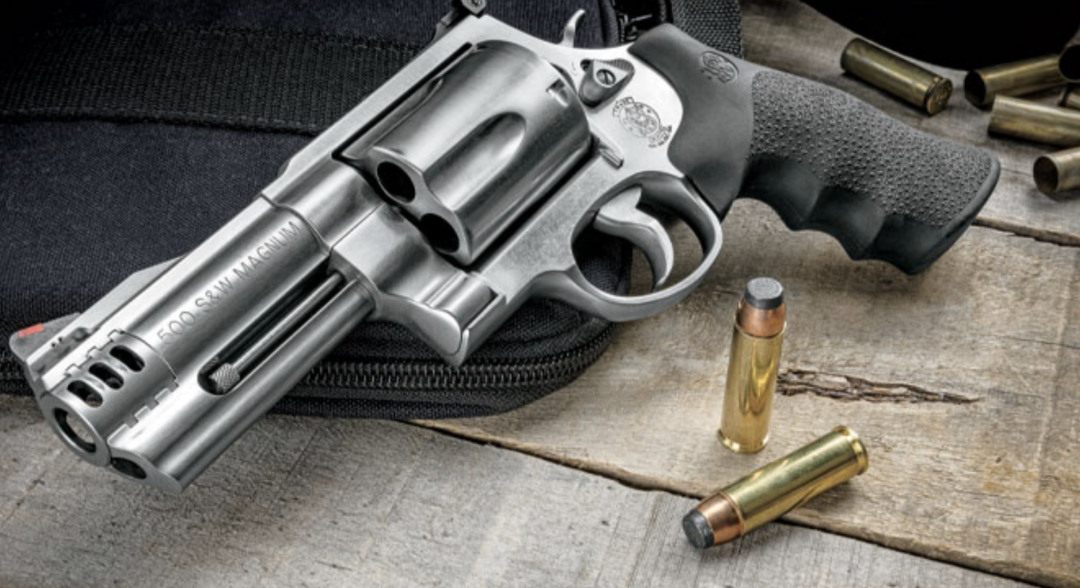 A silver revolver with a black grip is resting on a black case on a wooden surface, surrounded by several .500 S&W Magnum cartridges.